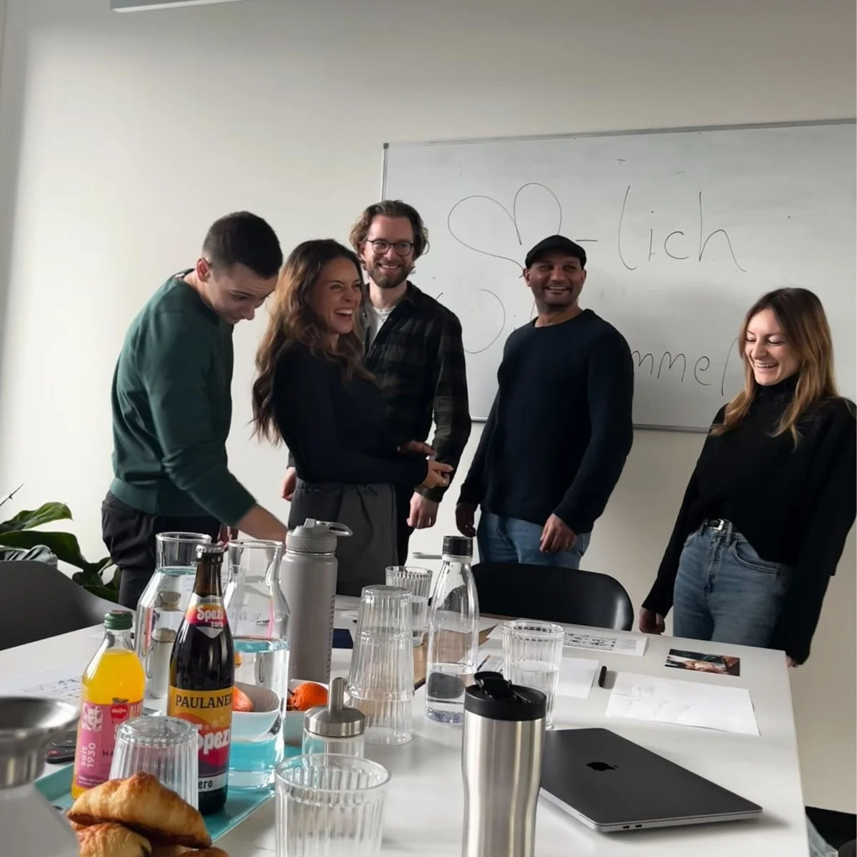 Fünf Personen lachen und stehen vor einer weißen Tafel in einem Büro, der Tisch im Vordergrund hat Getränke, Croissants und Unterlagen.