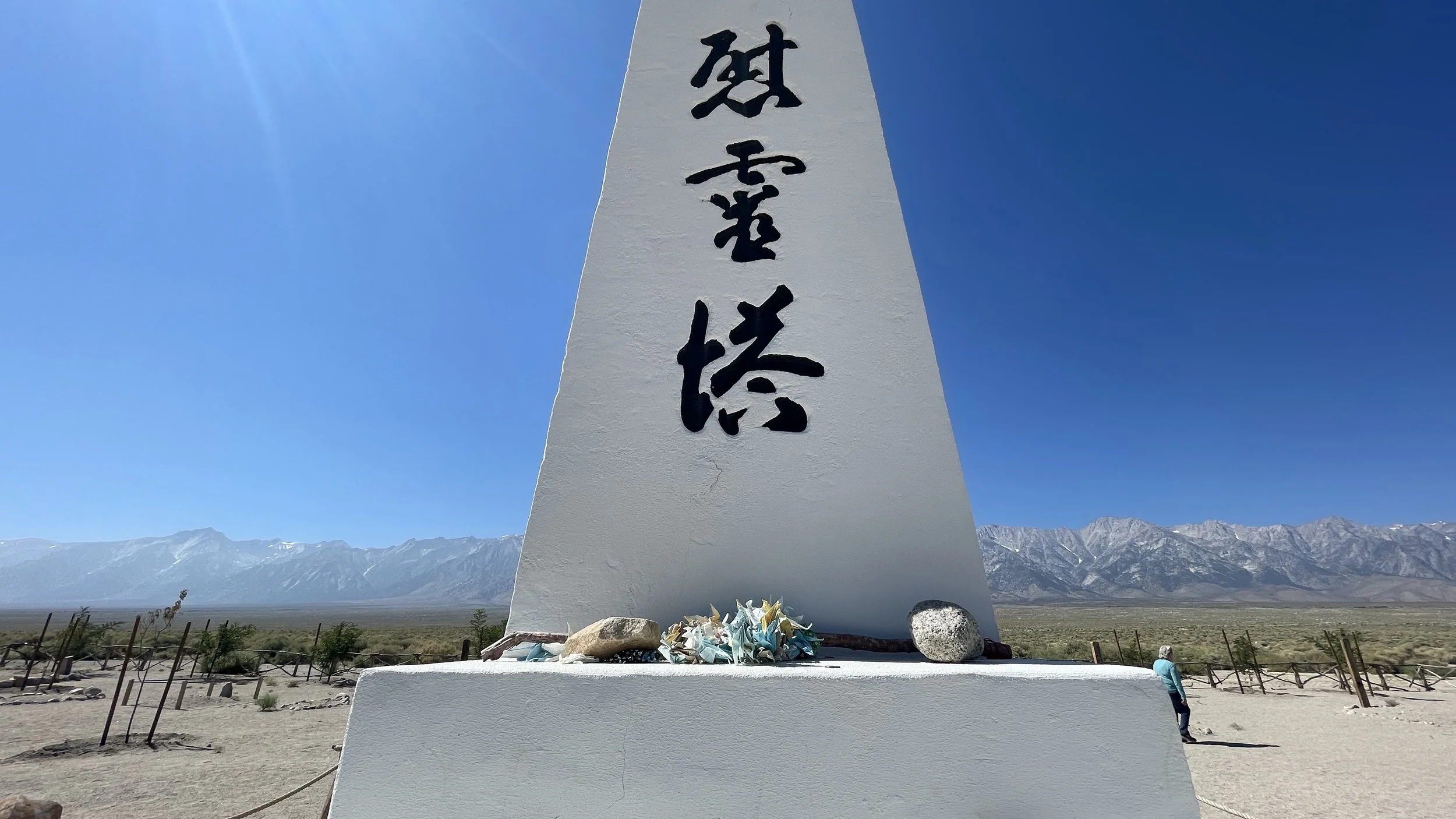 A white memorial obelisk with black Japanese characters, surrounded by rocks and flowers, at Manzanar National Historic Site in a vast desert landscape with mountains in the background under a clear blue sky.