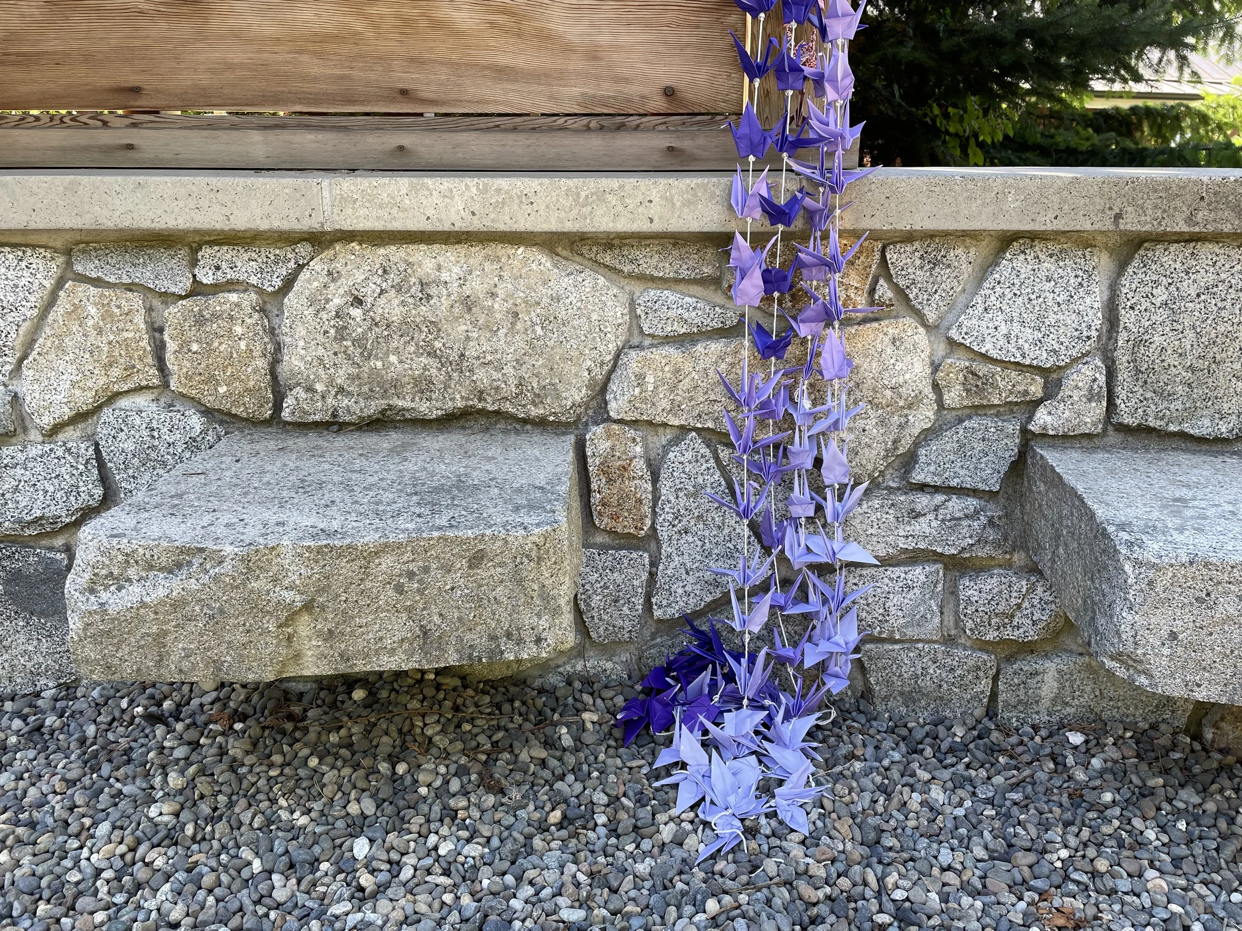 Purple and lavender origami cranes hanging in a vertical string decoration, positioned in front of a stone wall and gravel ground, with wooden and greenery background at the Japanese American Exclusion Memorial