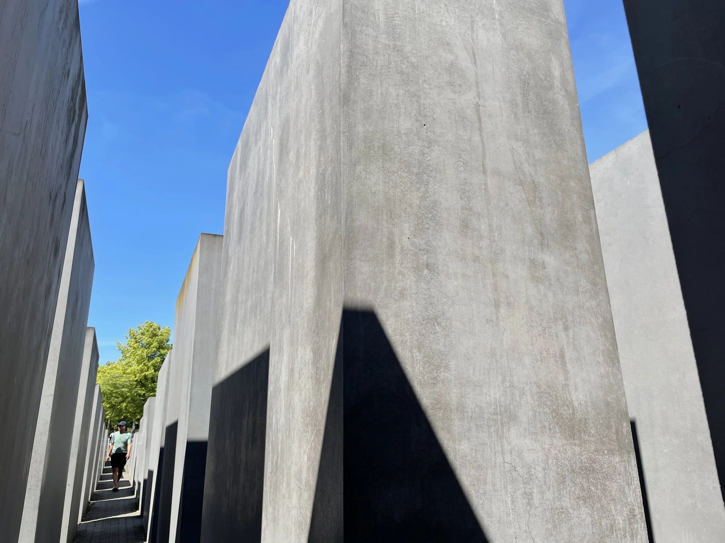 A person walking among concrete slabs at the Memorial to the Murdered Jews of Europe in Berlin, Germany, with a blue sky and green trees in the background.