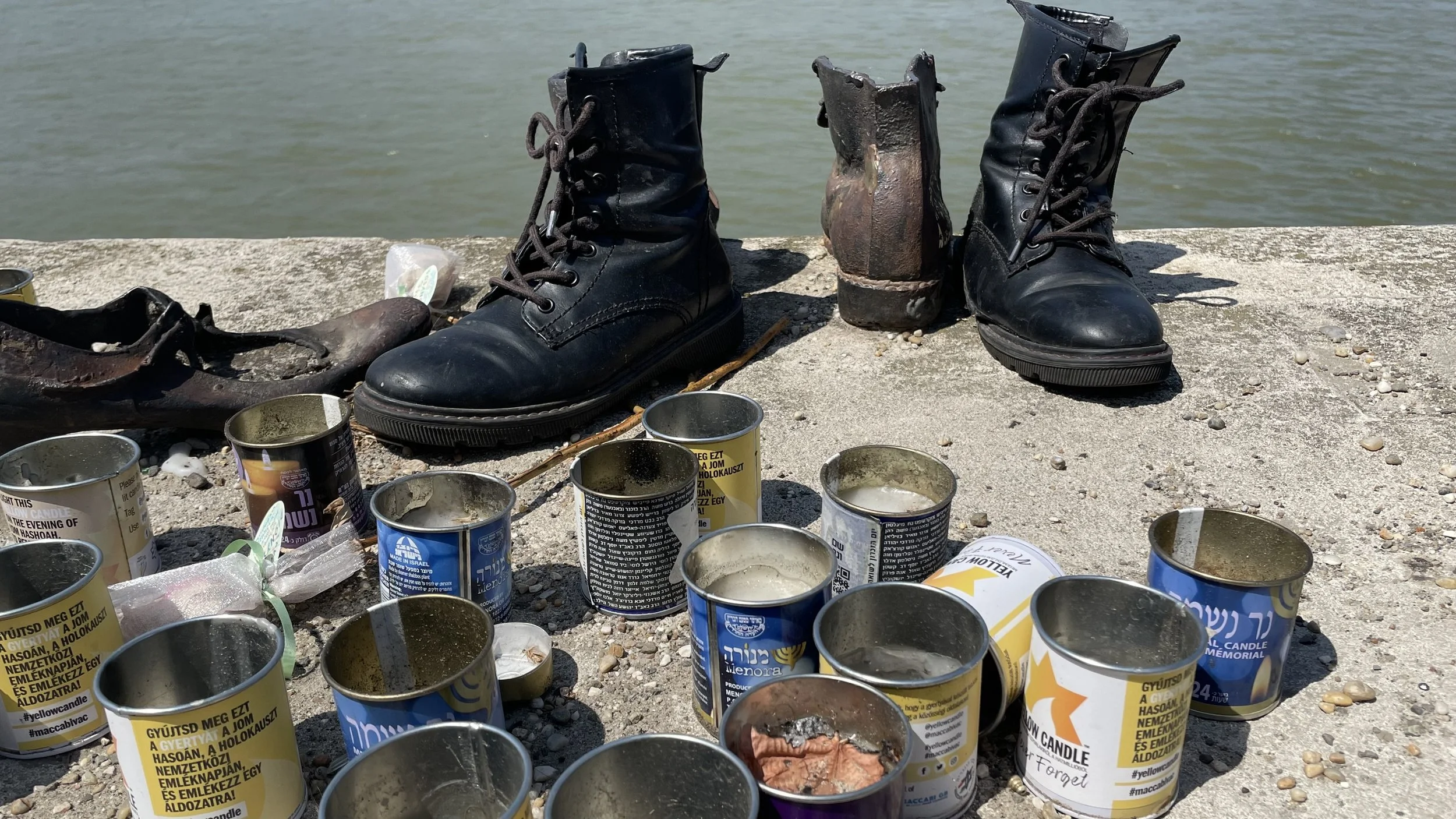A pair of black leather combat boots and a worn-out brown boot placed on a concrete surface near a body of water, with several candle tins scattered around them.