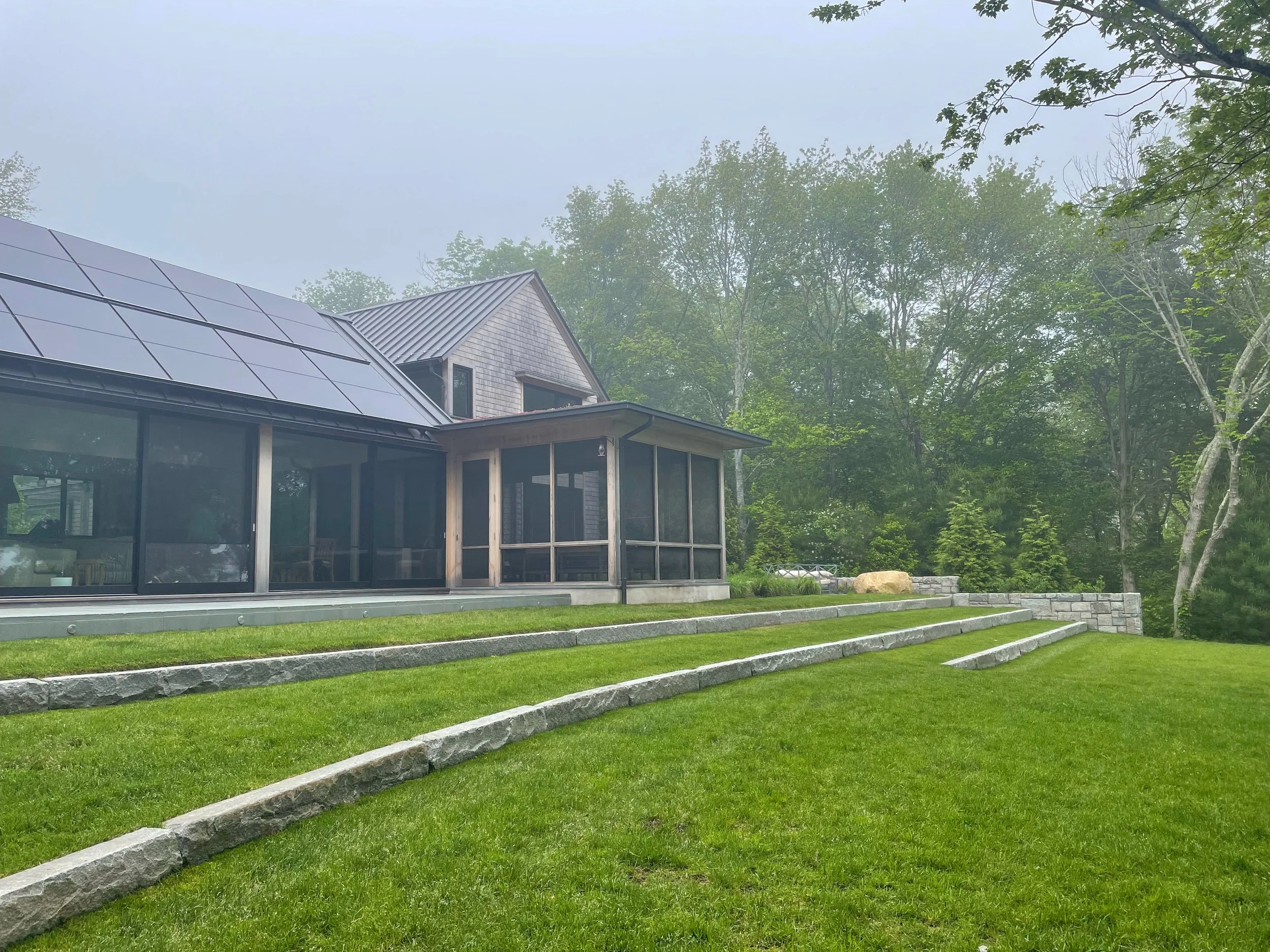 Modern house with solar panels on the roof, surrounded by green trees and a well-maintained lawn with stone steps.