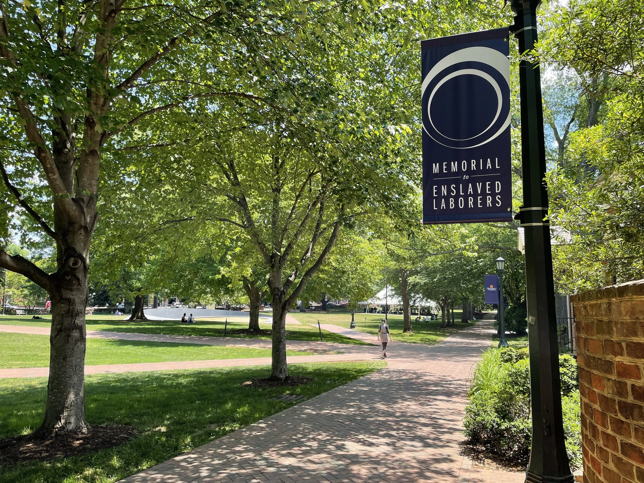 A park scene with green trees, a pathway, and a memorial banner that reads 'Memorial to Enslaved Laborers' hanging from a lamppost.