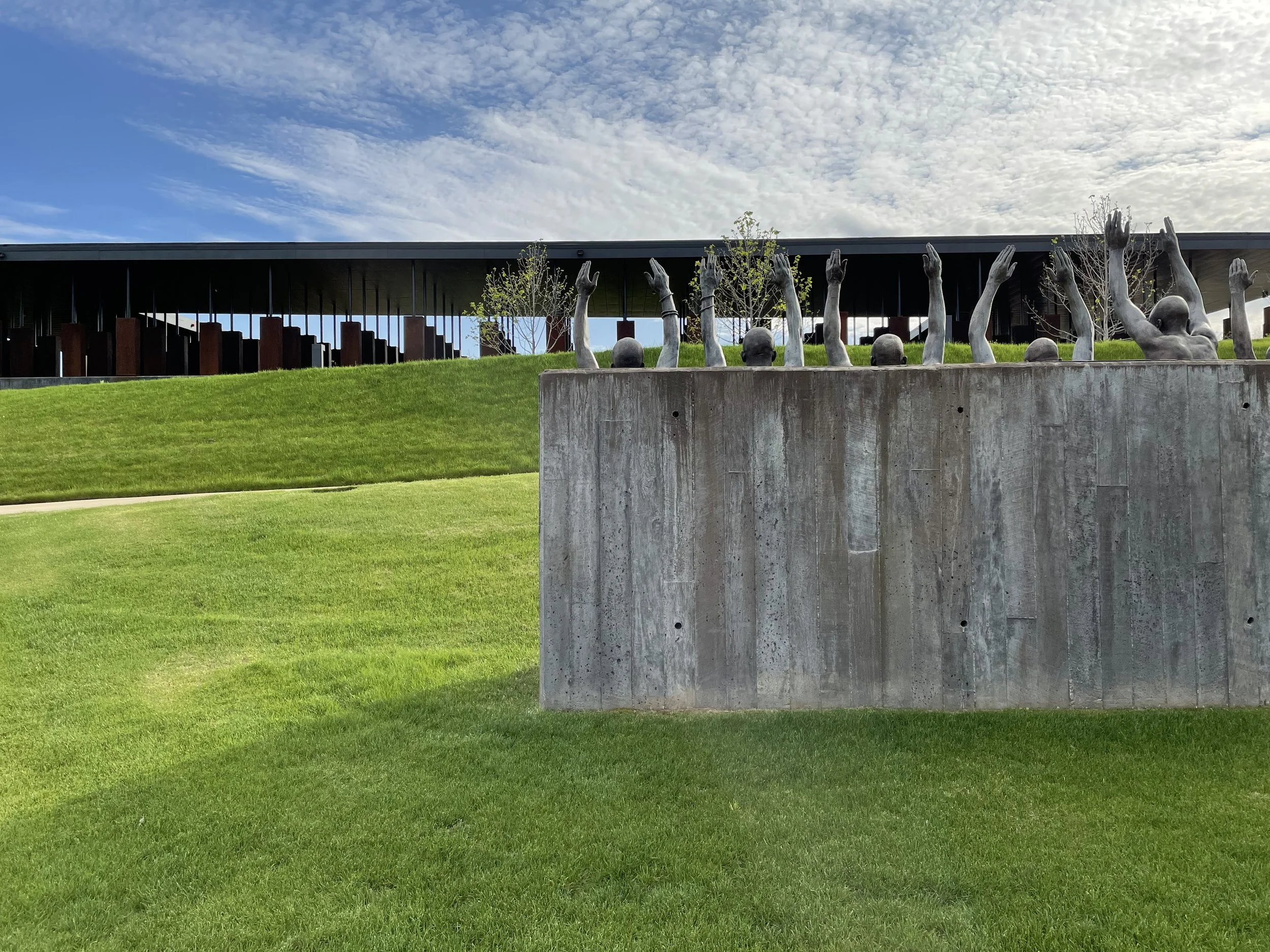 Outdoor sculpture by Hank Willis Thomas featuring multiple human arms and heads protruding from a concrete base, with a modern building and blue sky with clouds in the background.
