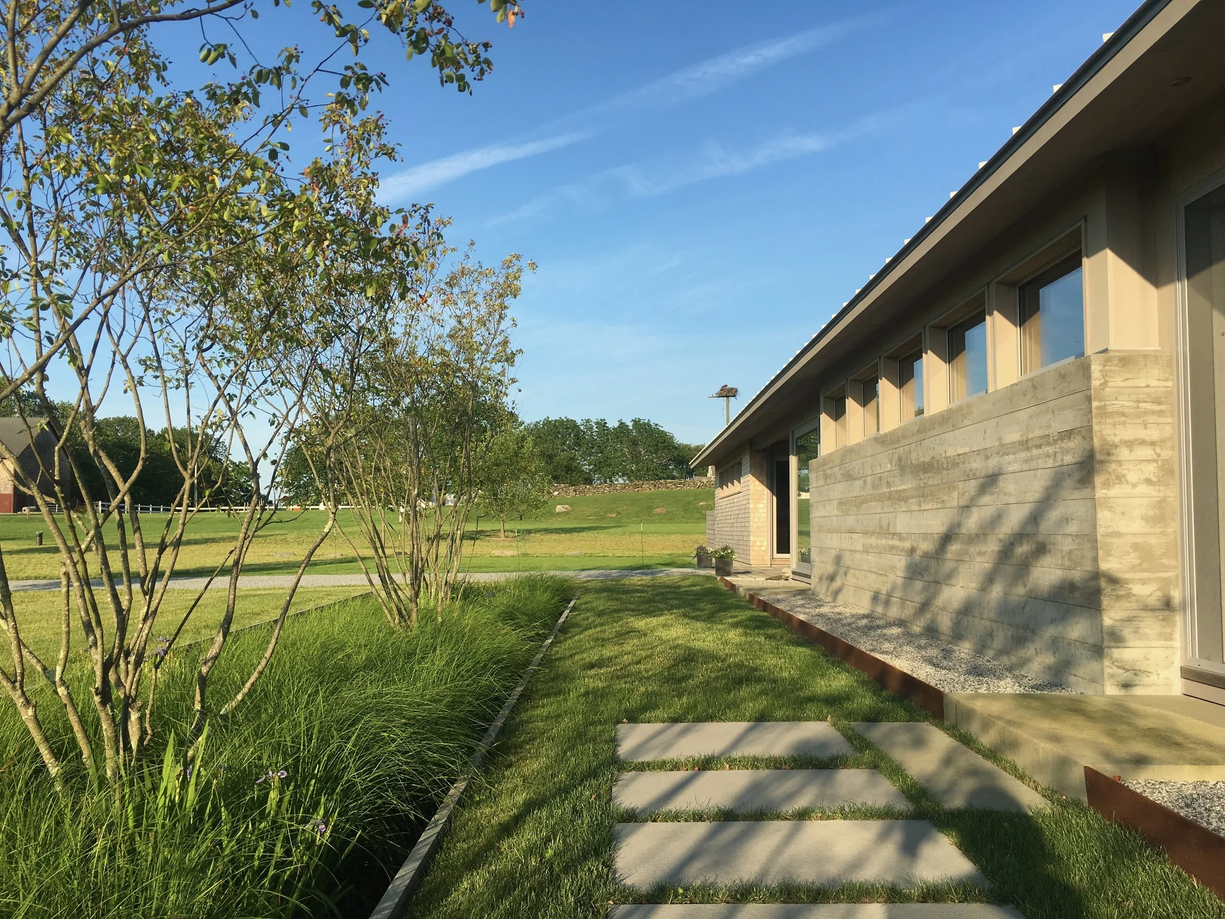 A modern house with large windows, a concrete exterior, and a sloped roof, surrounded by green grass, trees, and a clear blue sky.