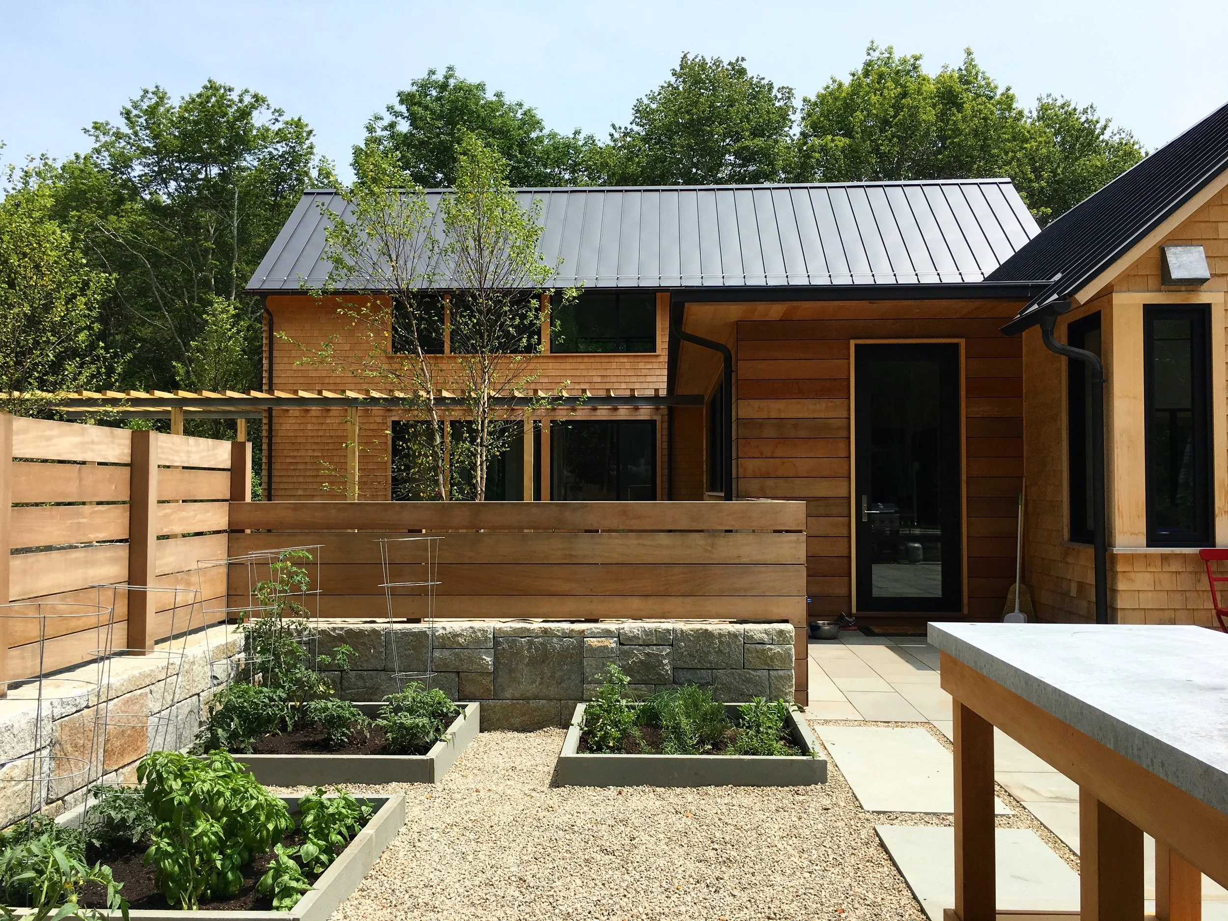 A backyard with raised garden beds containing green plants, a wooden privacy fence, a patio area, and a house with a metal roof and wooden siding surrounded by trees.