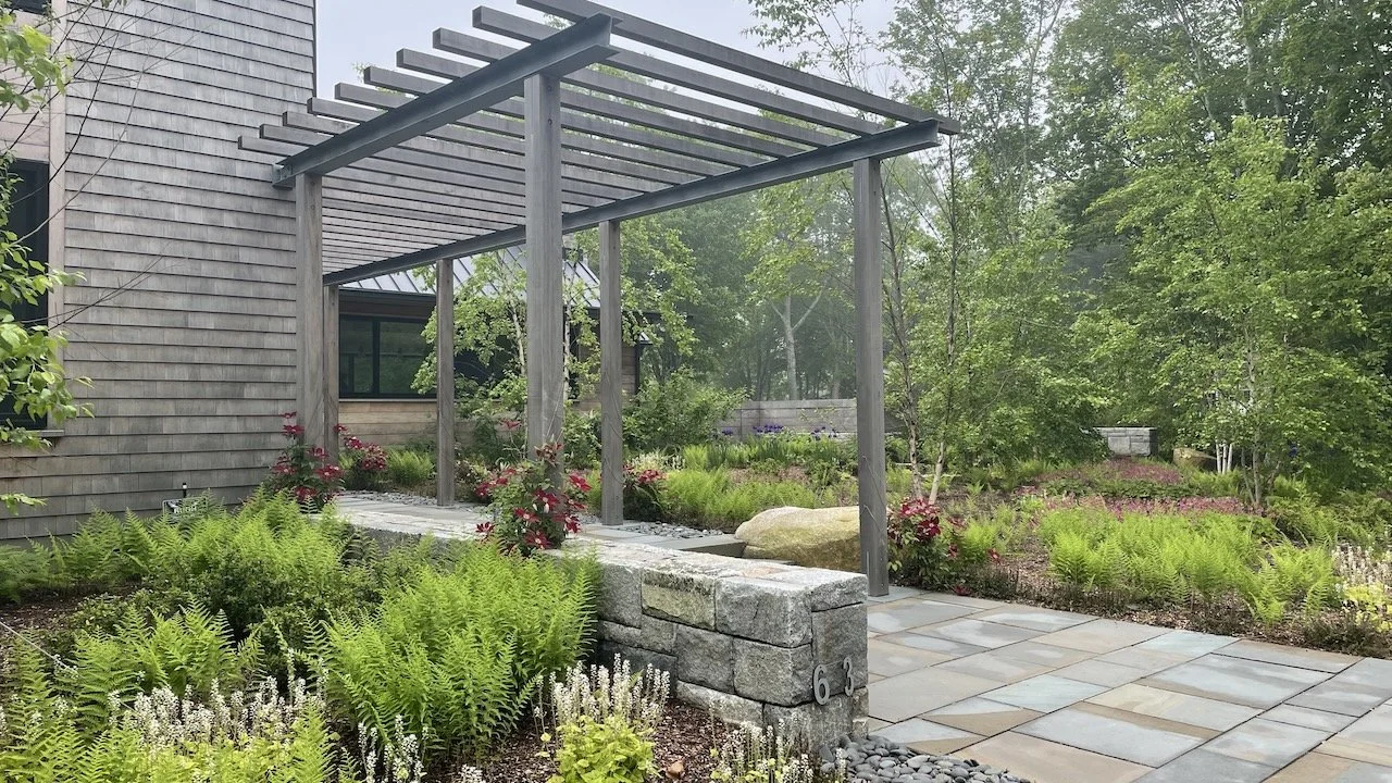 A landscaped backyard patio with a wooden pergola, stone paving, lush green plants, and trees.