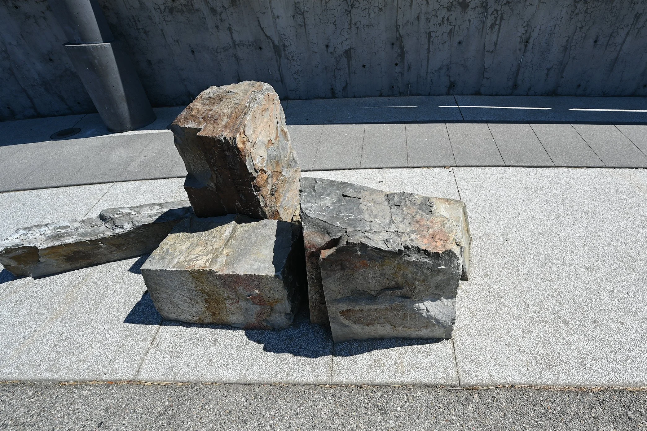 Multiple large rocks placed on a concrete surface in front of a concrete wall with shadows cast across the ground.