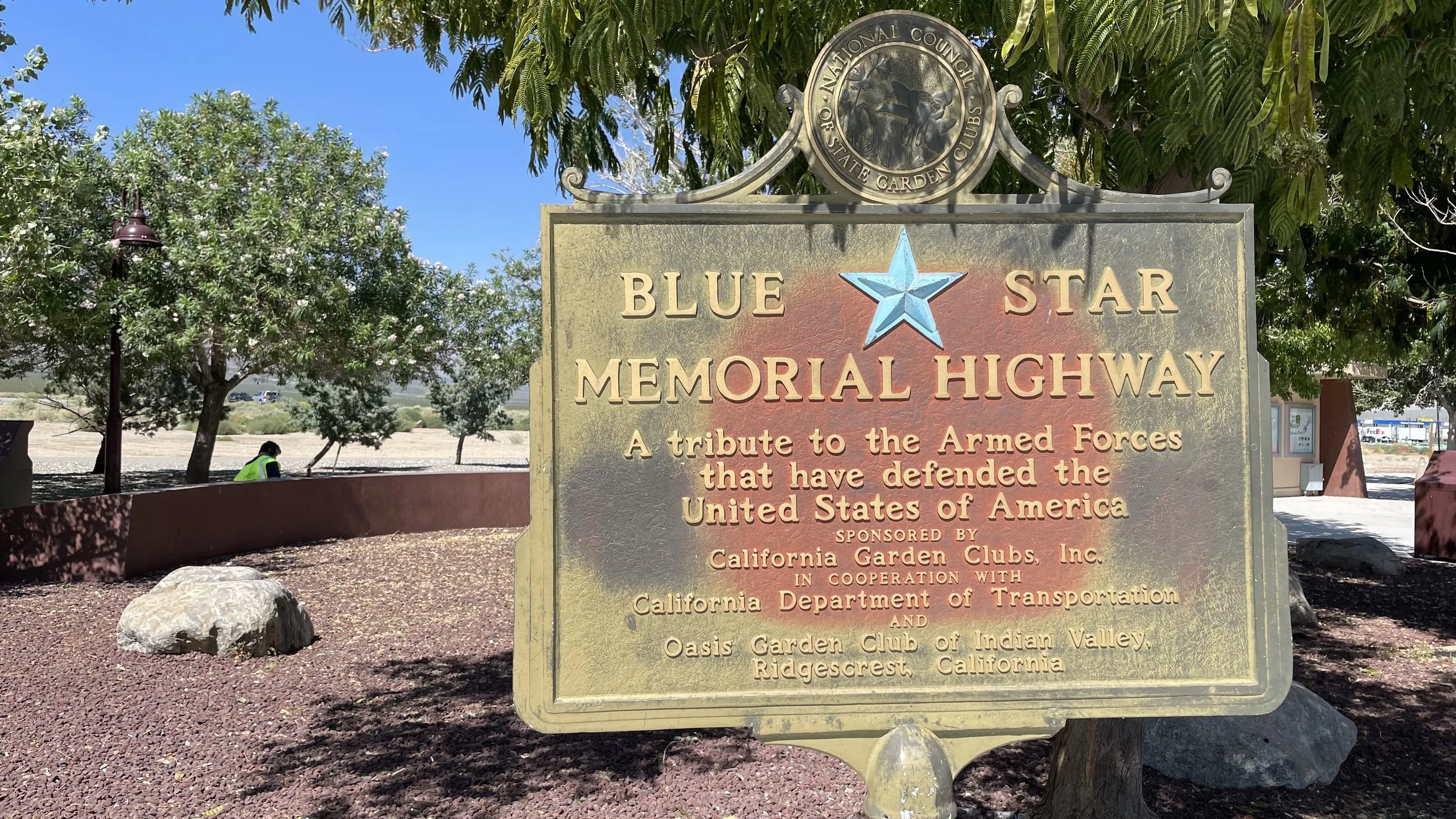 A metal sign for the Blue Star Memorial Highway, with a gold star in the center and a decorative top featuring a crest of the National Council of State Garden Clubs. The sign is situated in a park with trees, a person sitting on a bench, and a parkin