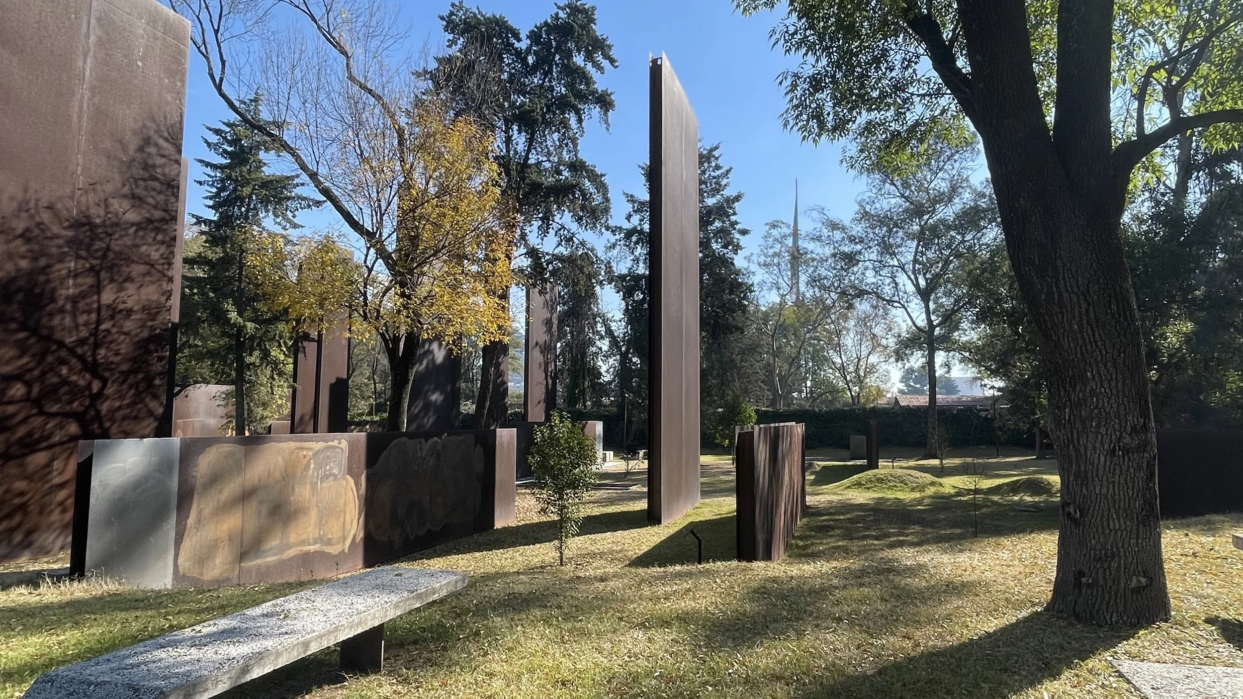 Outdoor memorial park with vertical metal slabs, trees, and a clear blue sky.