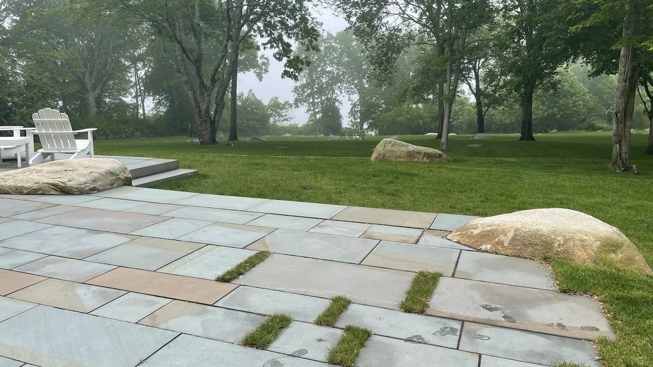 A backyard patio with paved stones and patches of grass, large rocks, and a lawn with trees in the background.