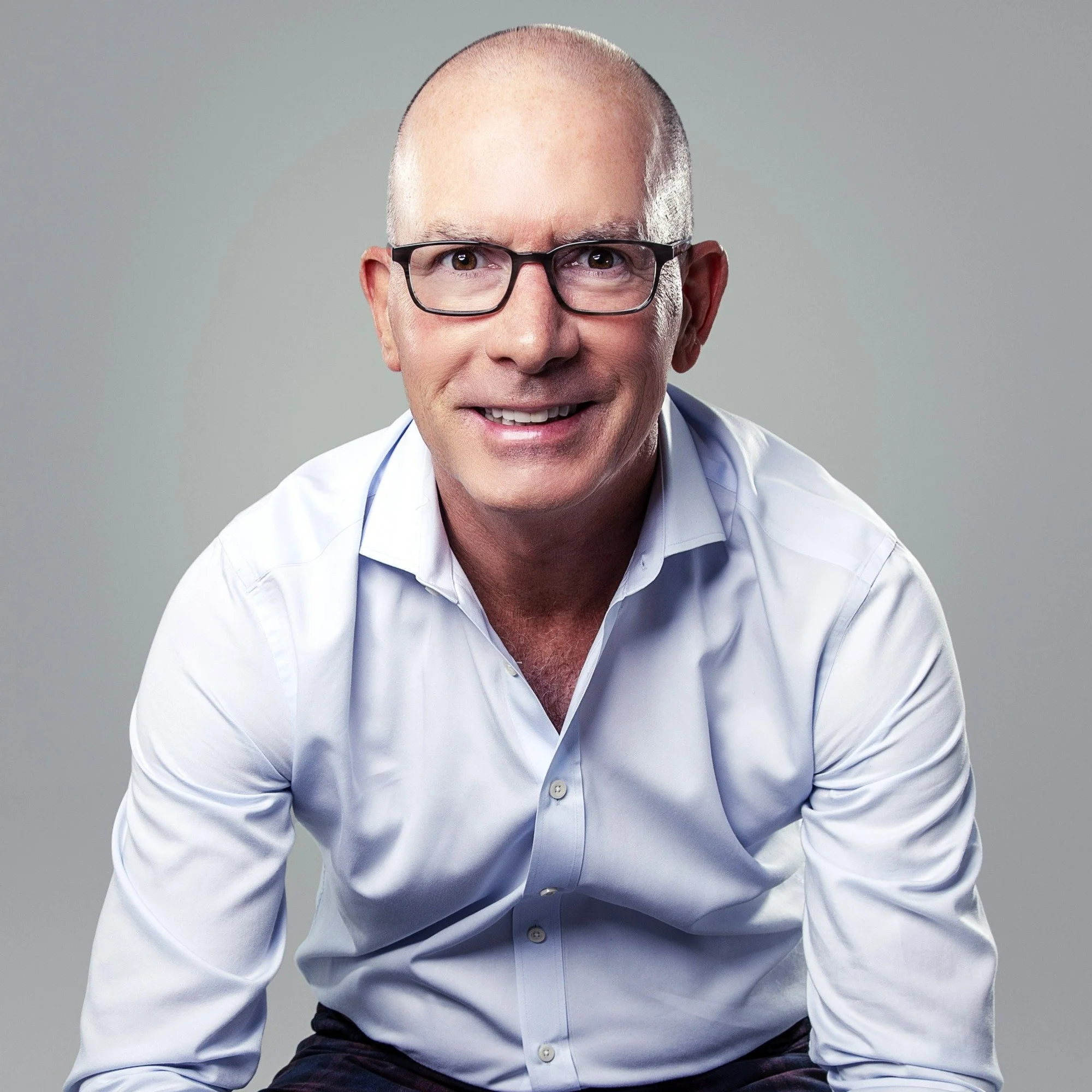 A middle-aged man with glasses, a bald head, wearing a white shirt, smiling at the camera against a neutral background.