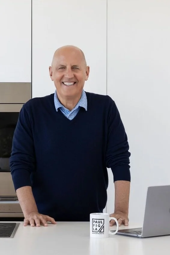 A smiling middle-aged man with a bald head, wearing a dark blue sweater over a light blue collared shirt, standing in a modern kitchen next to a white countertop with a laptop and a coffee mug that reads 'Paul Fig LA.'