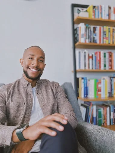 Charan Bashir. Compass certified coach. A man sitting on a gray couch in a room with a bookshelf filled with books behind him, smiling at the camera.