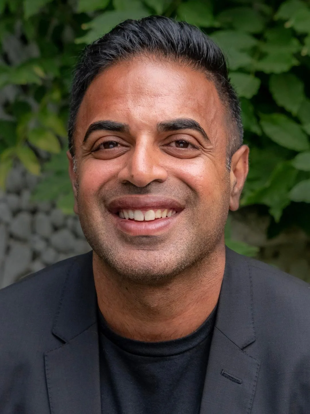 Chirag Shah. Compass certified coach. Close-up of a man with dark hair smiling, wearing a black blazer and shirt, with green foliage in the background.