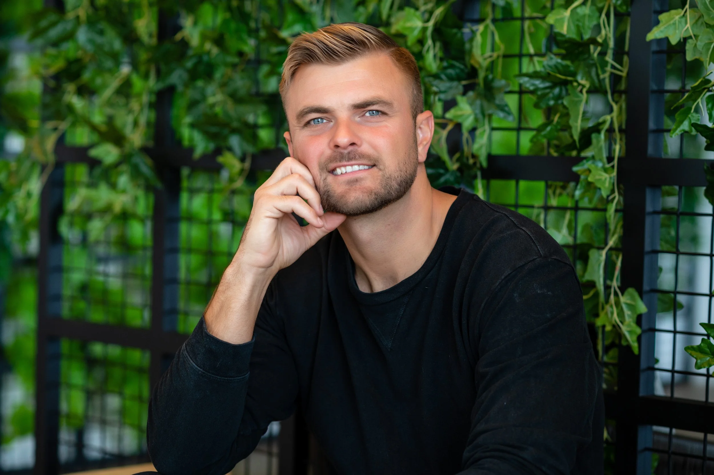 Jonathan Fox. Compass agent coach. A young man with blond hair, beard, and blue eyes, wearing a black long-sleeve shirt, sitting in front of a green leafy background, resting his chin on his hand and smiling at the camera.
