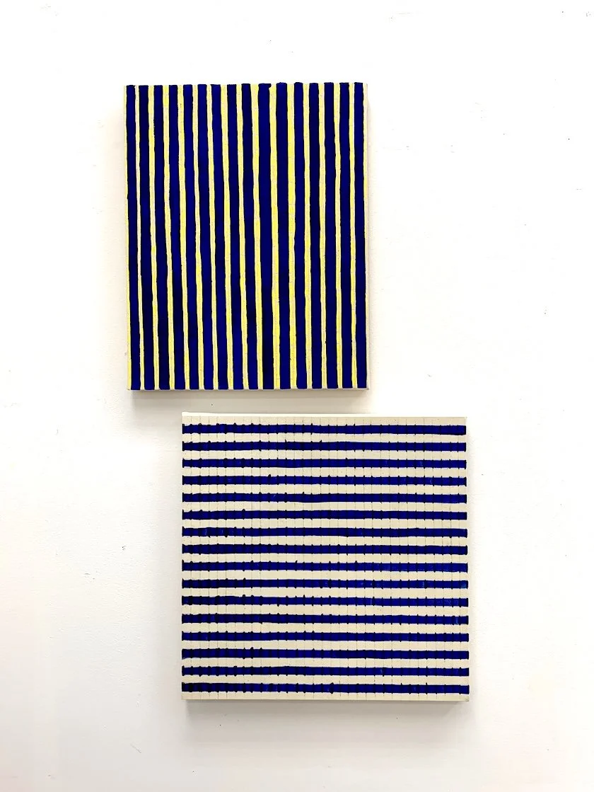 Two paintings with striped patterns, one yellow and blue vertical stripes, and the other blue and white horizontal stripes, hanging on a white wall.