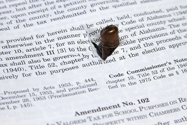 A bullet penetrating a page of legal or legal-style text, creating a hole around the impact area. The text includes references to amendments and code sections, suggesting a legal or legislative document.