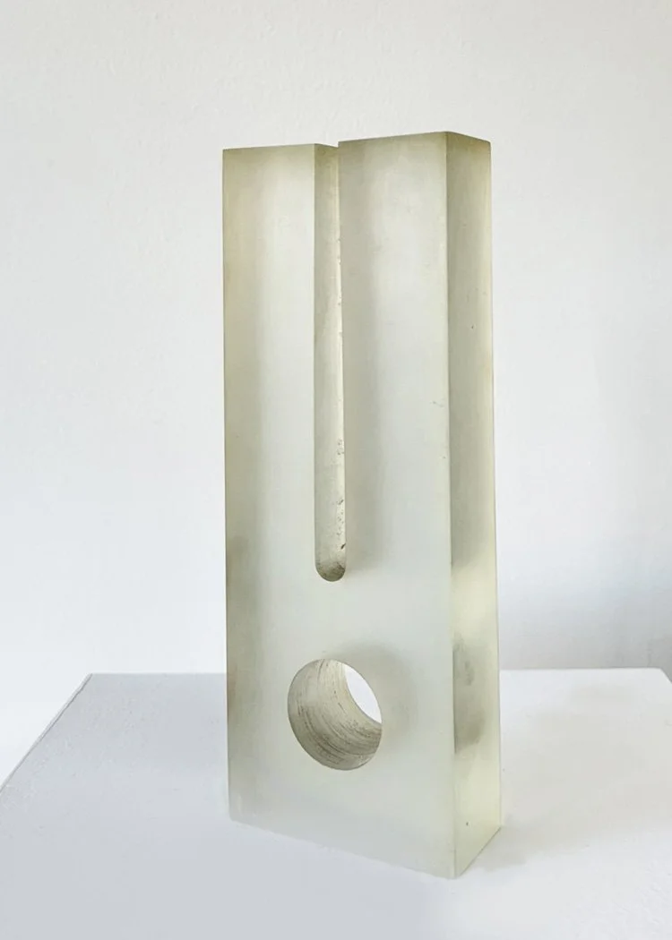 Abstract geometric acrylic sculpture by Angelo Granata with vertical slit and circular hole, made of translucent material, on display stand.