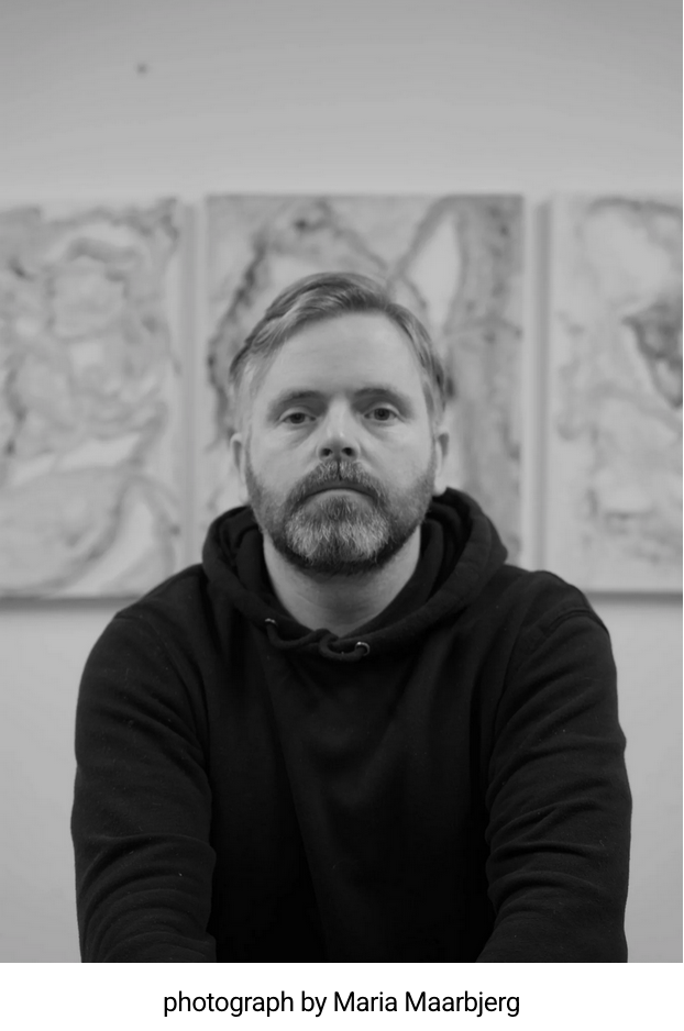 Black and white photo of a man with a beard and mustache, wearing a hoodie, sitting in front of three abstract paintings on the wall.