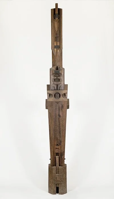 A tall, abstract wooden sculpture by Angelo Granata with angular shapes and carvings, against a plain background.