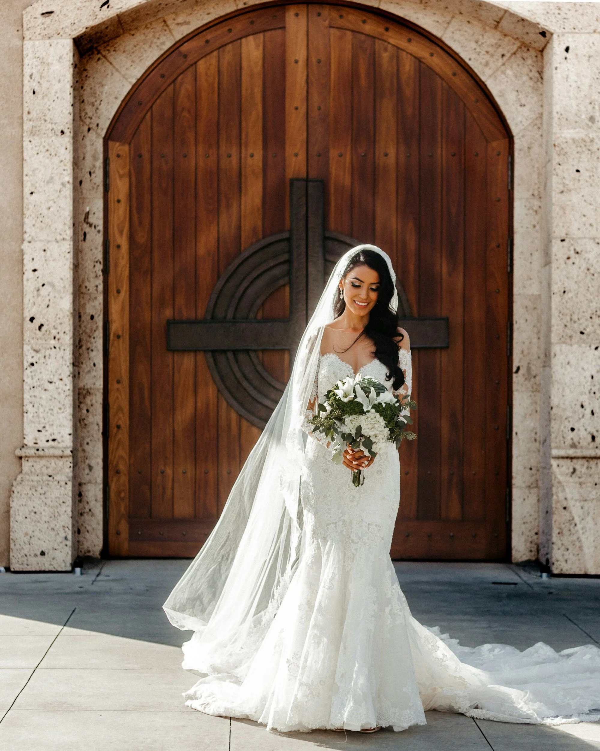 Bride in a white wedding dress holding a bouquet standing in front of large wooden door with circular design.