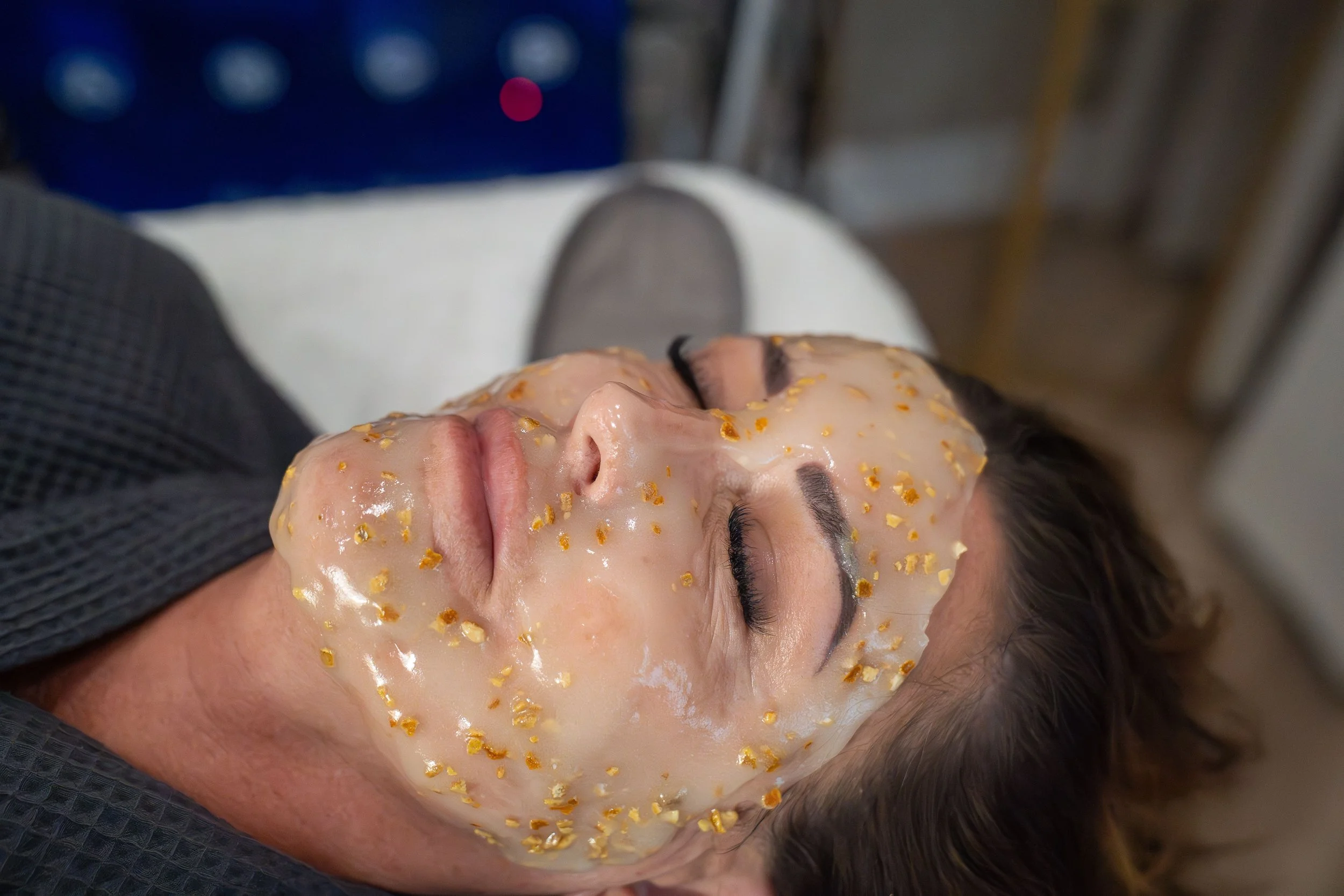 Woman lying down with facial mask containing orange peel bits, eyes closed, in a spa or skincare setting.