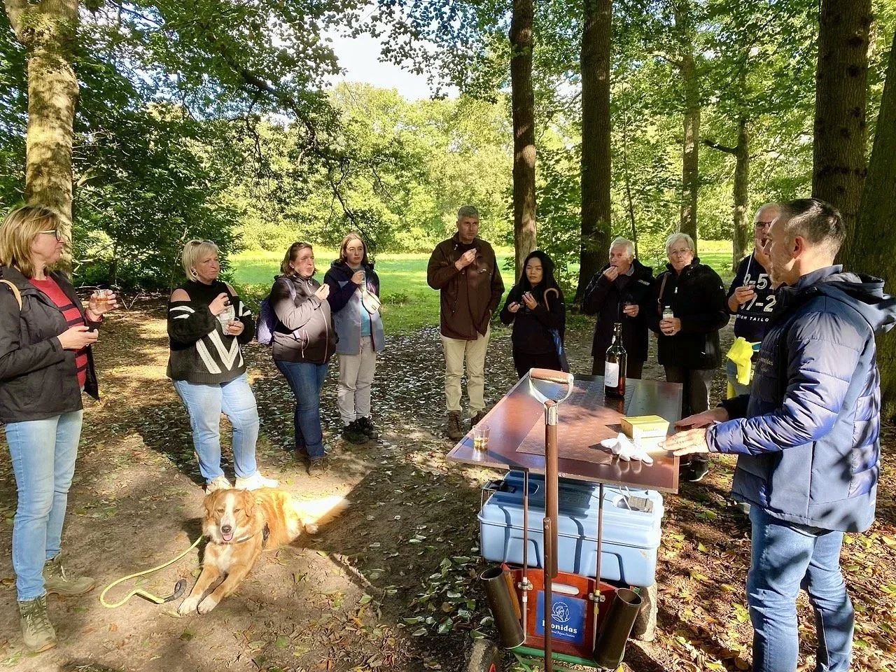 Groep mensen en een hond in een bos, staande rond een tafel met flessen en glazen.