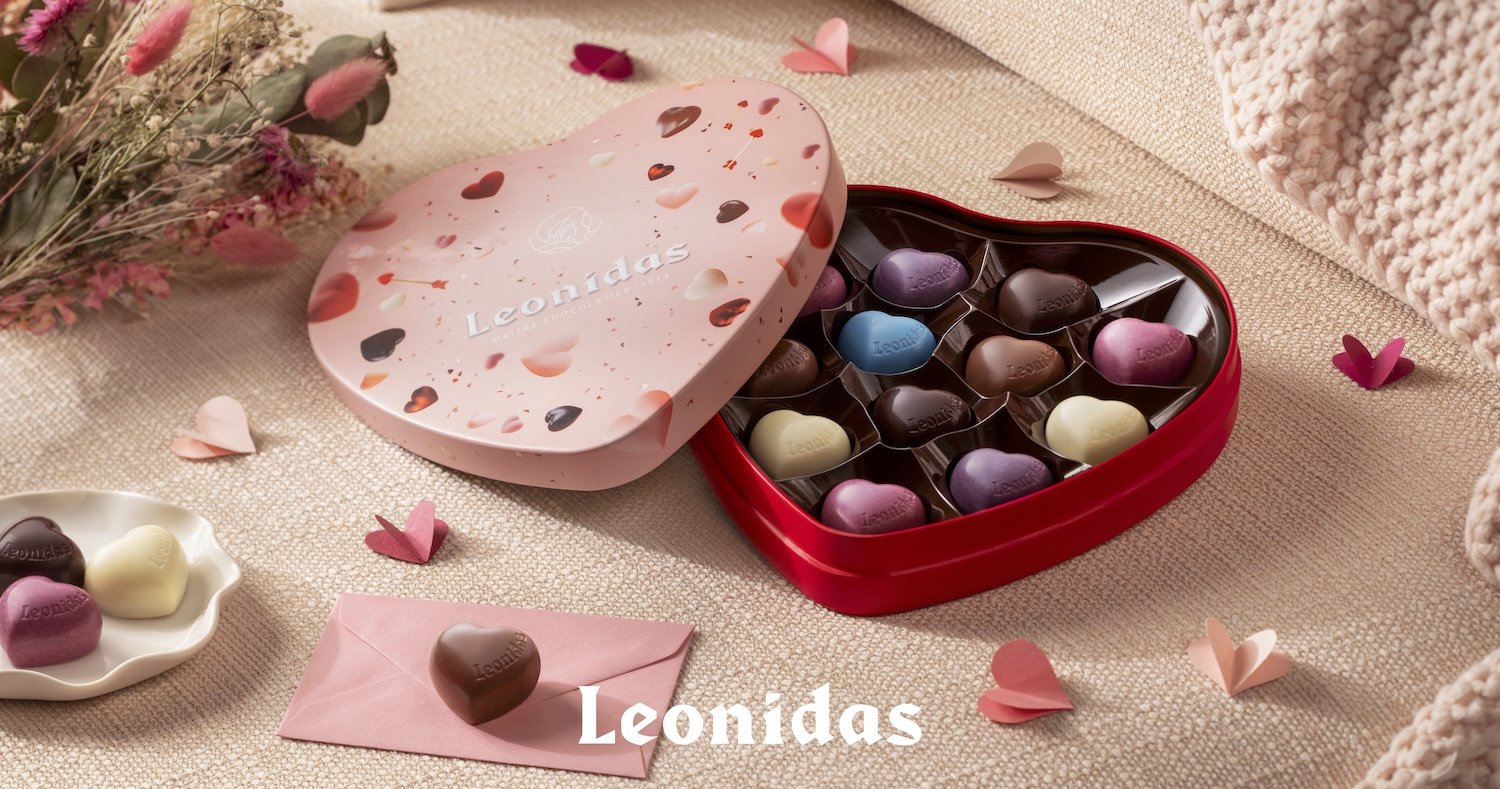 Een rood en roze hartvormig leodidas doosje met chocolaatjes hartjes, gelegen op een beige doek, omringd door papieren hartjes en bloemen.