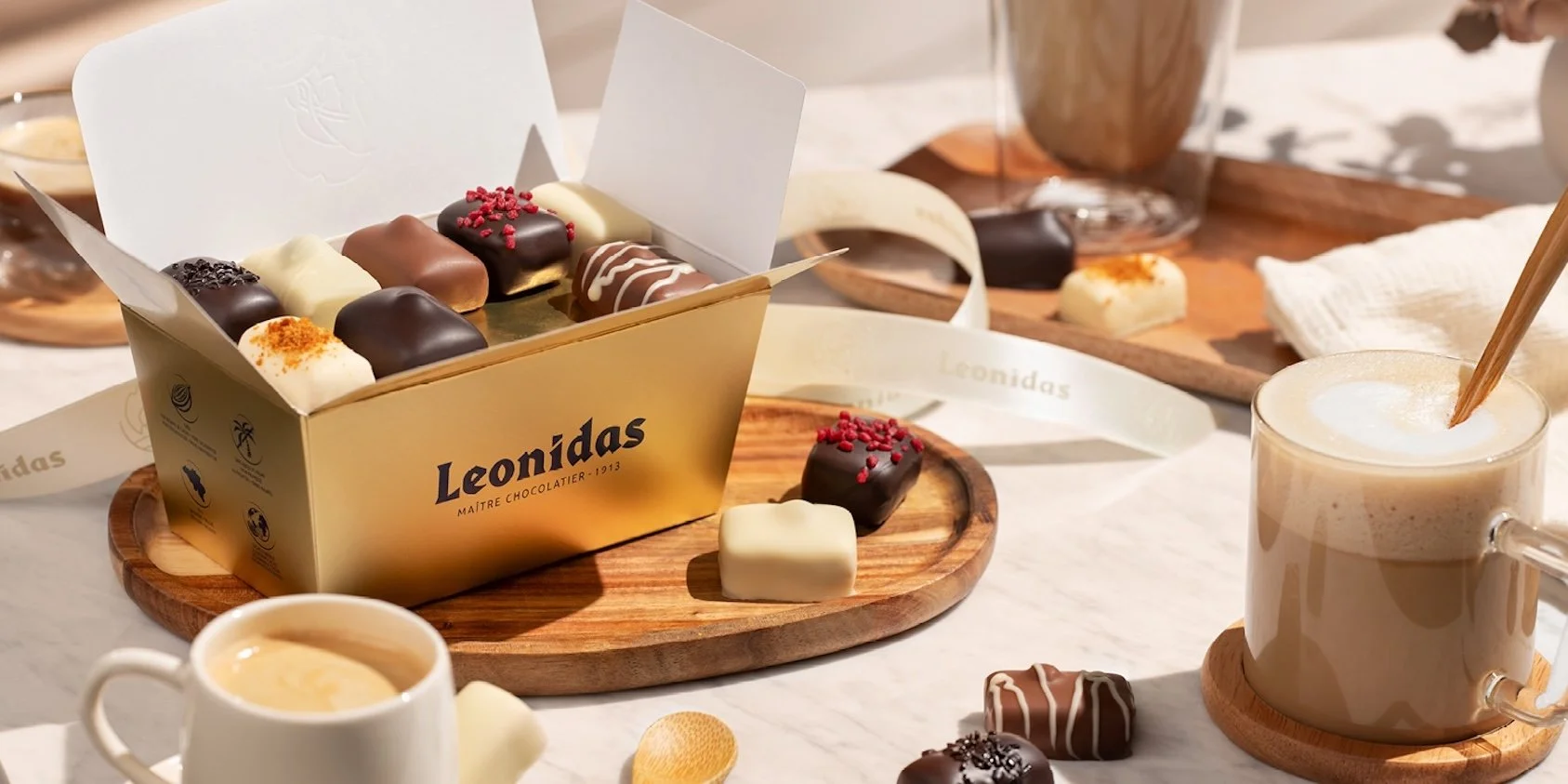 Een doos met verschillende manon pralines en bonbons op een houten bord, omgeven door kopjes koffie en thee op een witte tafel.