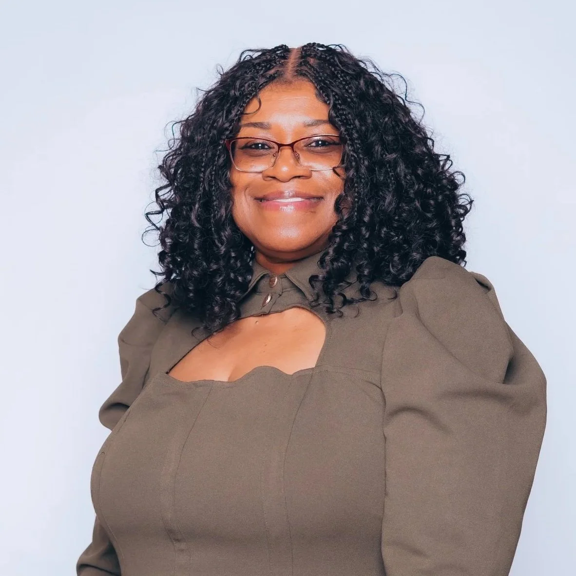 A woman with curly black hair, glasses, and a confident smile, dressed in a black blazer over a black and white striped top, standing with arms crossed against a plain beige background.