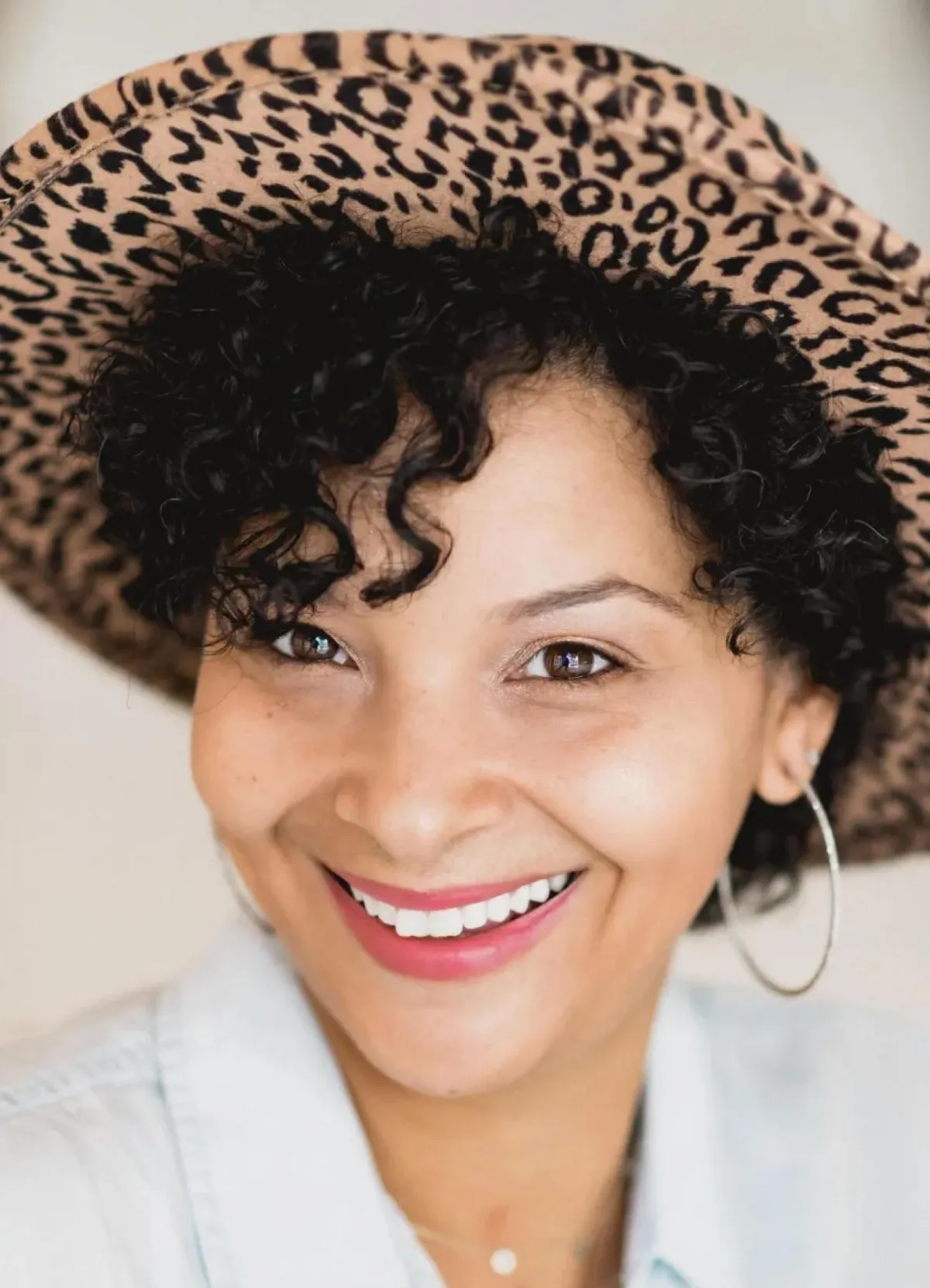 A woman with curly black hair, wearing a large leopard-print hat and hoop earrings, smiling at the camera.