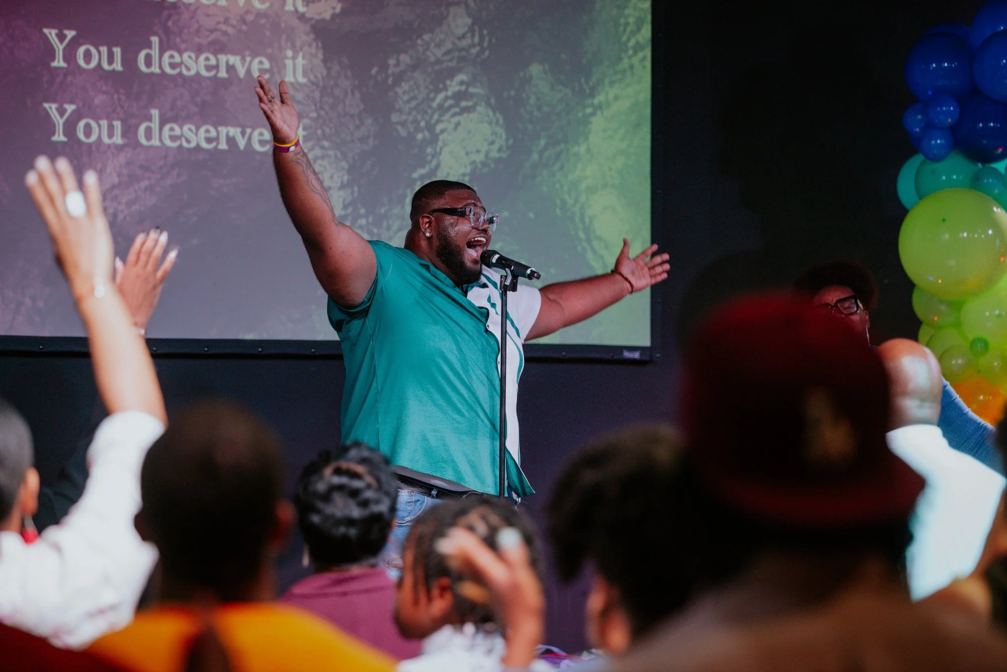 A man with glasses and a beard, wearing a teal shirt, stands with arms raised in front of a microphone on stage, leading a congregation in worship. The audience is raising their hands. A large screen behind displays the words "You deserve it" twice. Colorful balloons are on the right side of the stage.