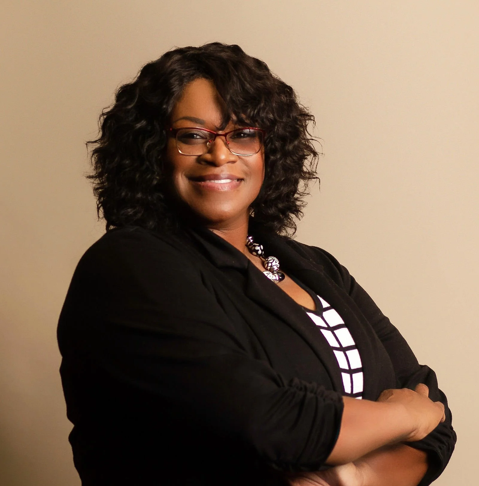 A woman with curly black hair, glasses, and a confident smile, dressed in a black blazer over a black and white striped top, standing with arms crossed against a plain beige background.