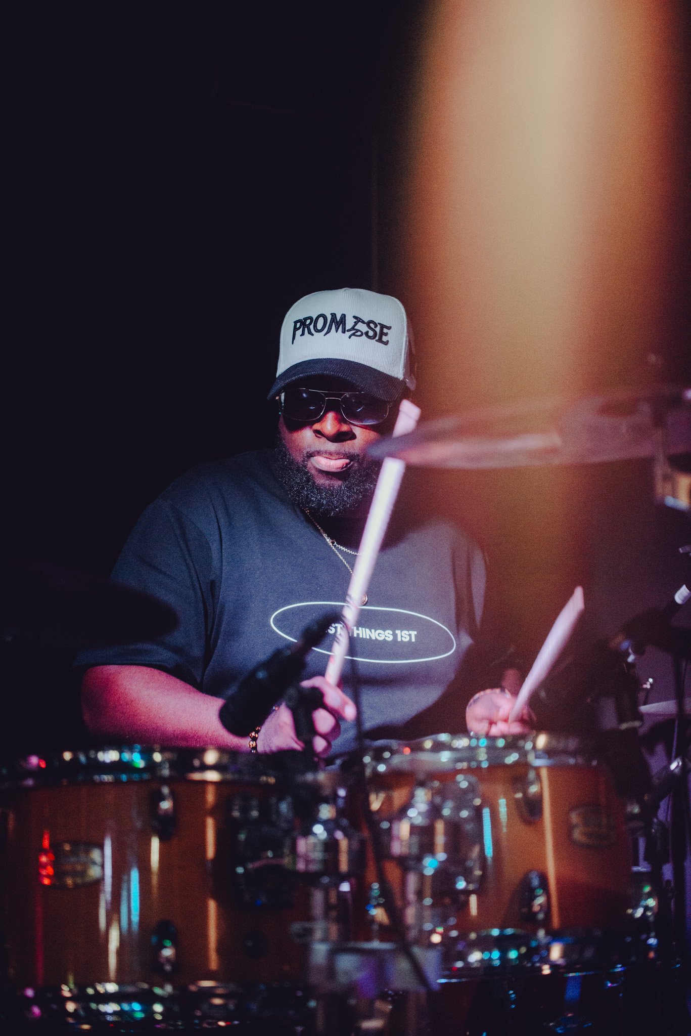 A man wearing sunglasses, a gray cap with 'PROM 3SE' on it, and a black T-shirt with the phrase 'DOING THINGS 1ST' is playing drums in a dimly lit setting.