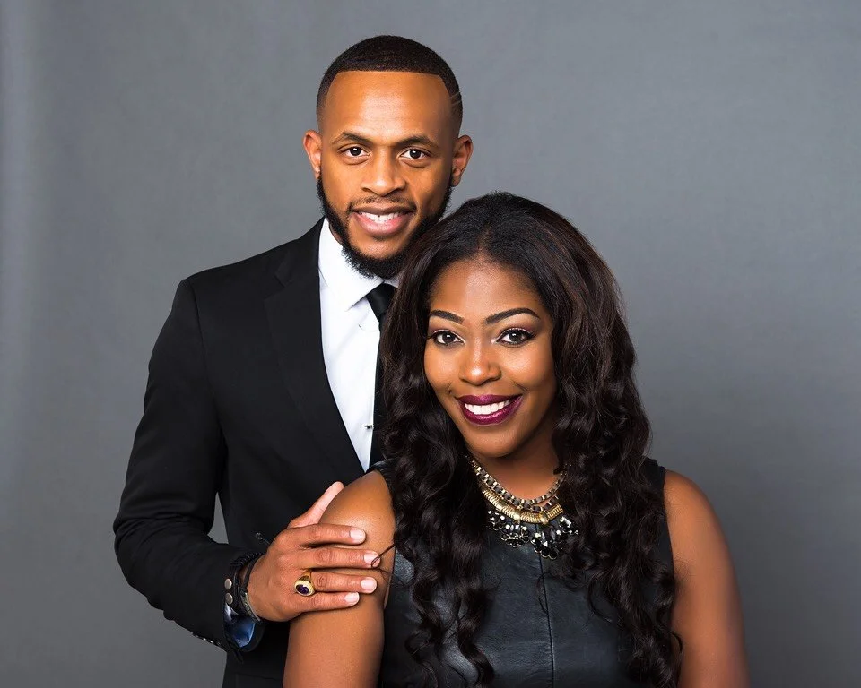 A well-dressed man in a black suit and white shirt standing behind a woman with long wavy black hair, wearing a black sleeveless top and colorful jewelry, both smiling against a gray background.
