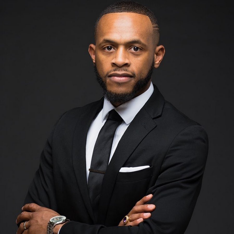 A professional man wearing a black suit, white shirt, and black tie, posing with arms crossed against a dark background.