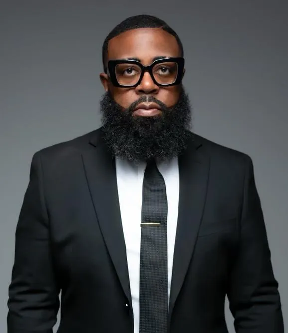 Man with glasses, full beard, wearing a black suit, white shirt, and black tie, against a gray background.