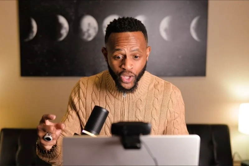 A man with a beard and short curly hair, wearing a tan cable knit sweater, looking surprised at a laptop. Behind him is artwork of moon phases and a lamp on a side table.