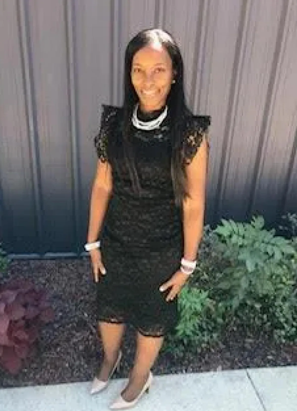 A woman wearing a black lace dress and white high heels, standing outdoors in front of a gray fence and green plants.