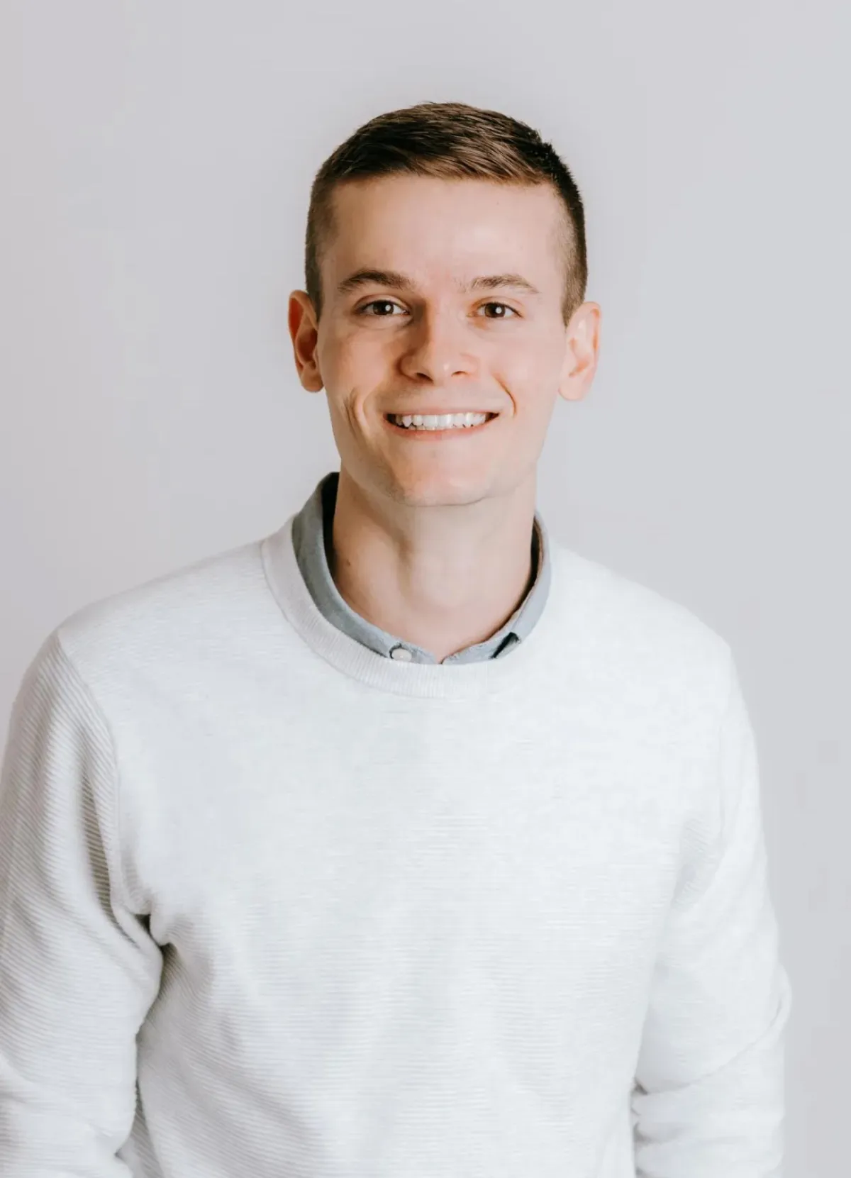 A young man with short brown hair, smiling, wearing a light-colored sweater over a collared shirt, standing against a plain light gray background.