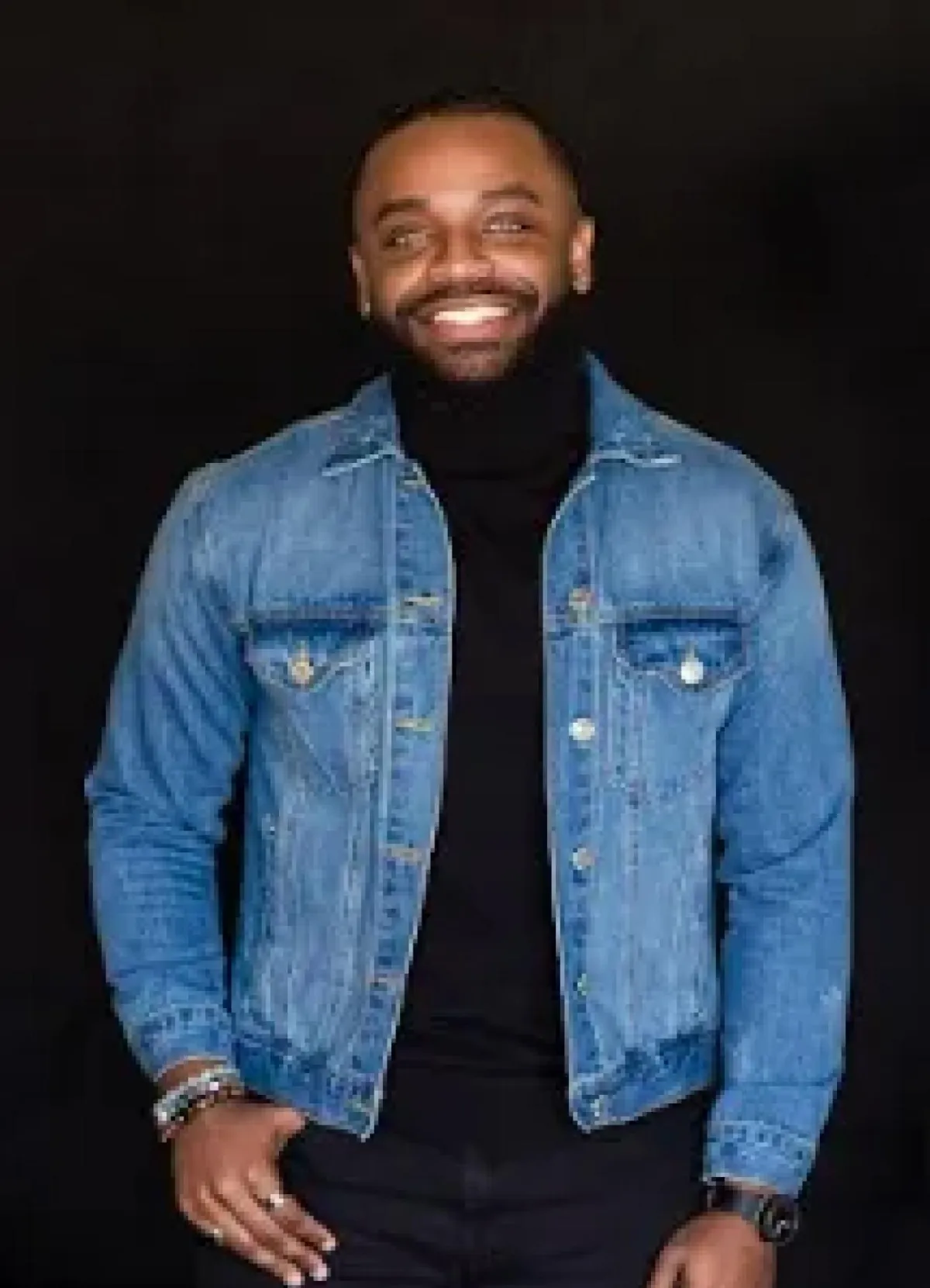 A smiling man with a beard wearing a denim jacket over a black turtleneck, standing against a black background.