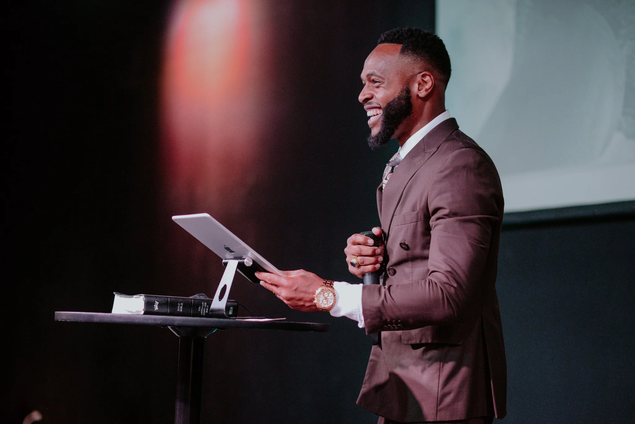 A man in a brown suit with a watch and rings, smiling, holding a microphone, standing near a tablet on a stand, giving a presentation in a dark room.