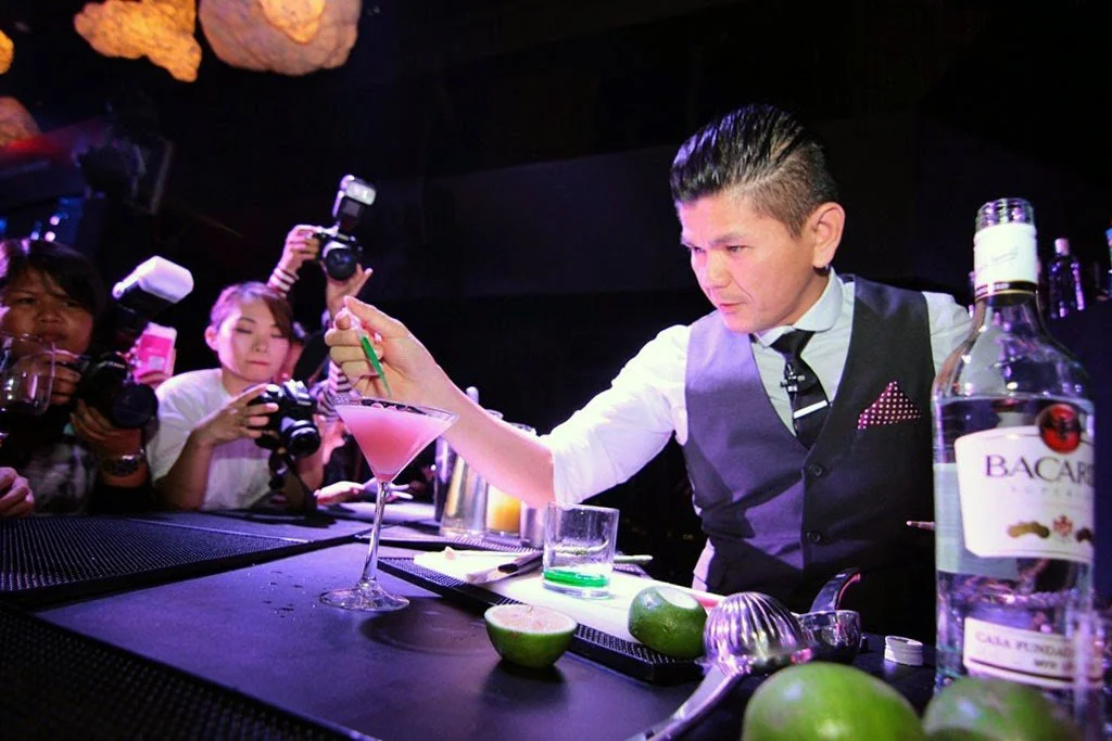 Bartender in a vest preparing a pink cocktail at a bar while photographers and onlookers watch.