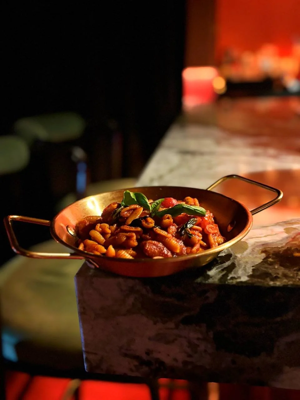 Close-up of a copper dish filled with pasta, topped with green herbs, on a marble countertop in a dimly lit restaurant.