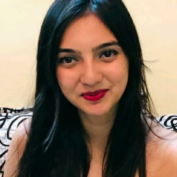 A woman with long black hair, wearing red lipstick, smiling at the camera, seated against a plain light-colored wall.