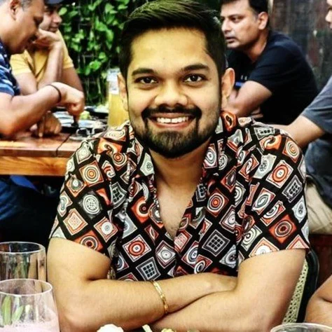 A man with a beard and mustache smiling at the camera, wearing a patterned shirt, sitting at a table in a restaurant or cafe with other people in the background.