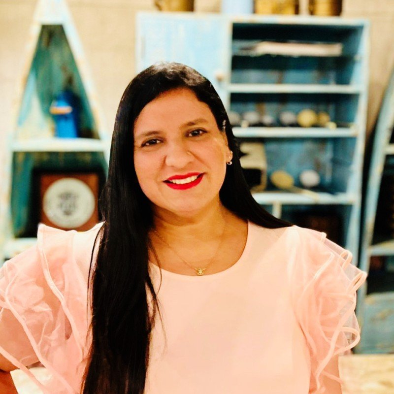 A woman with long dark hair, wearing a light pink ruffled blouse and red lipstick, smiling in front of a wooden bookshelf with decorative items.
