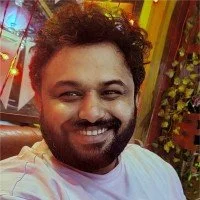 A man with curly hair and a beard smiling, wearing a white shirt, in an indoor setting with colorful decorations and plants in the background.
