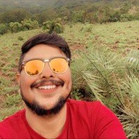 A smiling man with a beard and sunglasses taking a selfie outdoors in a grassy area with trees in the background.